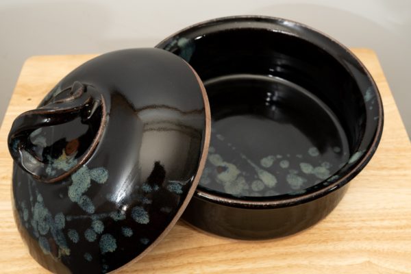 the photograph shows a clay coyote bread baker glazed in midnight garden with its lid resting on the left side of the base. this offers the viewer a look into the bread baker and shows the midnight garden pattern continues inside the bread baker. the bread baker and lid are resting on a small light colored wooden table. the background is a white wall. the photograph is lit with white light.