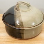 the photograph shows a clay coyote bread baker glazed in mint chip resting on a small light colored wooden table. the table is in front of a white wall. the bread baker has its lid on. the photograph is lit with white light.