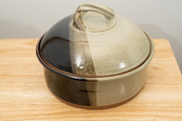 the photograph shows a clay coyote bread baker glazed in mint chip resting on a small light colored wooden table. the table is in front of a white wall. the bread baker has its lid on. the photograph is lit with white light.