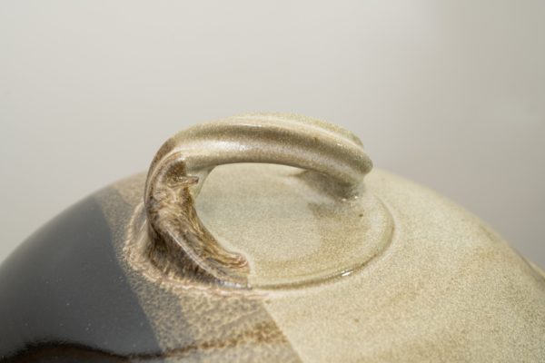 the photograph shows a close up view of the top of a clay coyote bread baker lid. the focus of the photograph is the twisted handle on the top of the break bakers lid. the background is white. the photograph is lit with white light.