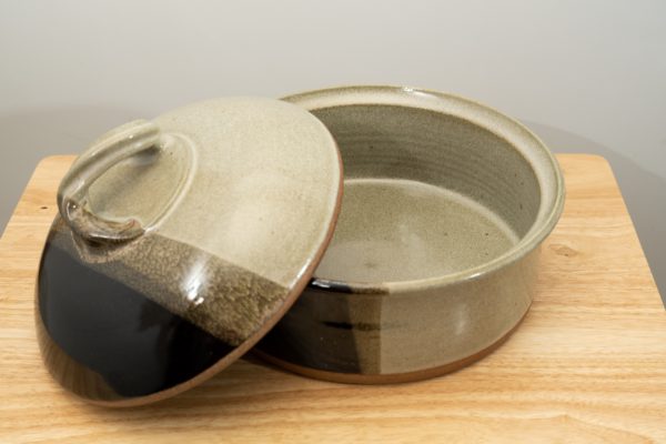 the photograph shows a clay coyote bread baker glazed in mint chip with its lid resting on the left side of the base. this offers the viewer a look into the bread baker. the bread baker and lid are resting on a small light colored wooden table. the background is a white wall. the photograph is lit with white light.