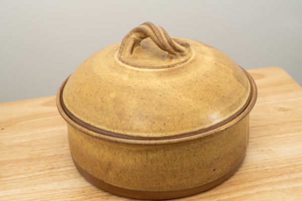 the photograph shows a clay coyote bread baker glazed in yellow salt resting on a small light colored wooden table. the table is in front of a white wall. the bread baker has its lid on. the photograph is lit with white light.