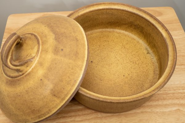 the photograph shows a clay coyote bread baker glazed in yellow salt with its lid resting on the left side of the base. this offers the viewer a look into the bread baker. the bread baker and lid are resting on a small light colored wooden table. the background is a white wall. the photograph is lit with white light.