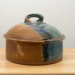 the photograph shows a clay coyote bread baker glazed in joes blue resting on a small light colored wooden table. the table is in front of a white wall. the bread baker has its lid on. the photograph is lit with white light.