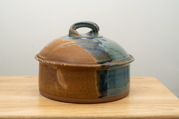 the photograph shows a clay coyote bread baker glazed in joes blue resting on a small light colored wooden table. the table is in front of a white wall. the bread baker has its lid on. the photograph is lit with white light.