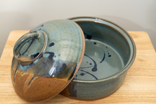 the photograph shows a clay coyote bread baker glazed in joes blue with its lid resting on the left side of the base. this offers the viewer a look into the bread baker, and shows how the joes blue pattern continues inside the bread baker. the bread baker and lid are resting on a small light colored wooden table. the background is a white wall. the photograph is lit with white light.