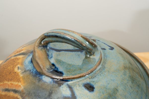 the photograph shows a close up view of the top of a clay coyote bread baker lid. the focus of the photograph is the twisted handle on the top of the break bakers lid. the background is white. the photograph is lit with white light.