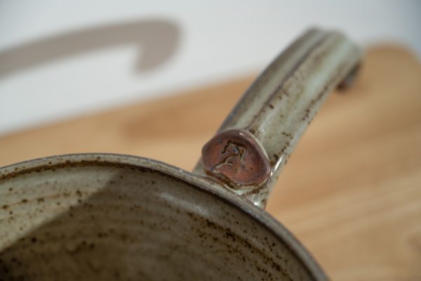 the photograph shows a close up of a clay coyote flameware medium saucepans handle, and the little coyote stamp detail. the handle is pointed towards the upper right corner. the background is a white wall. the pan is resting on a out of focus light colored wooden table. the background is a white wall.