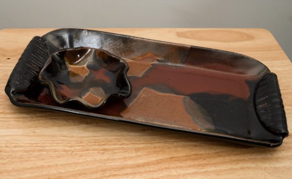 the photograph shows a clay coyote large tray and little dipper combo resting on a wooden table. the large tray and little dipper are glazed in merlot. the small table they are resting on is light colored wood. the tray is at a slight angle on the table, with the left side pointed towards the upper left corner of the photograph and the right side pointed towards the lower right corner. the little dipper is the circular or star style. the little dipper is on the left side of the large tray. the background is a white painted wall and is barely visible on the very upper edge of the photograph and the upper right corner.