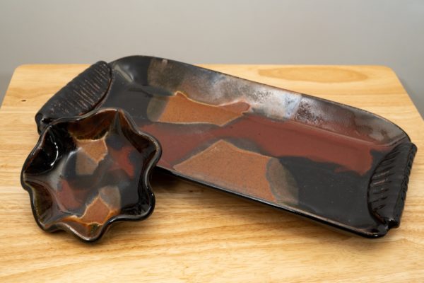 the photograph shows a clay coyote large tray and little dipper combination resting on a light colored wooden table. the large tray and little dipper are both glazed in merlot. the little dipper is the star or circular style. the little dipper is propped up against the edge of the large tray, slightly elevating it, and offering a clear view of the merlot design on the inside of the little dipper. the tray is resting at an angle . the left side of the tray is pointed towards the upper left corner of the photograph, while the right side is pointed towards the lower right corner. the background is a white painted wall and is just visible on the upper edge of the photograph.