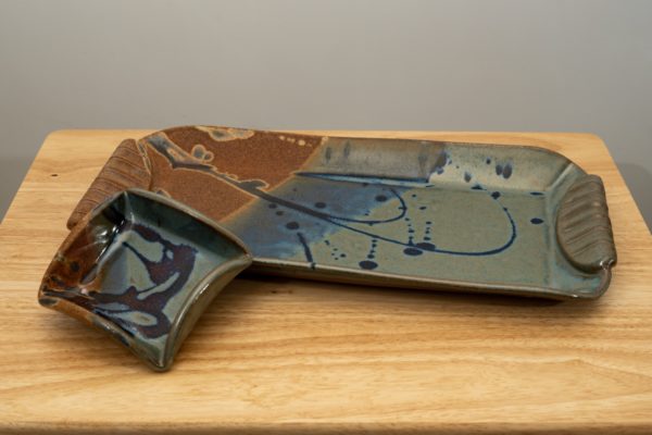 the photograph shows a clay coyote large tray and little dipper resting on a light colored wood table. the large tray and the little dipper are glazed in joes blue. the little dipper is resting on the left side of the tray. the little dipper is outside of the large tray, but is propped against the lip of it to stand it up a little bit. the little dipper is the square style of dipper. the section of brown on the glaze for both pieces is facing the left side of the photograph. the background is a white painted wall.