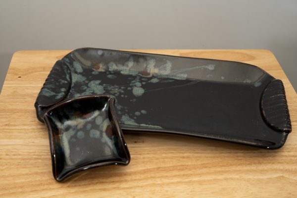 the photograph shows a clay coyote large tray and little dipper resting on a light colored wooden table. the large tray and the little dipper are both glazed in midnight garden. the little dipper is resting outside of the large tray, but propped up against the trays lip, to angle the little dipper slightly. the little dipper is resting on the left side of the tray. the large tray is angled slightly with the left side pointed towards the upper left corner and the right side pointed slightly to the lower right. the background is a white wall.