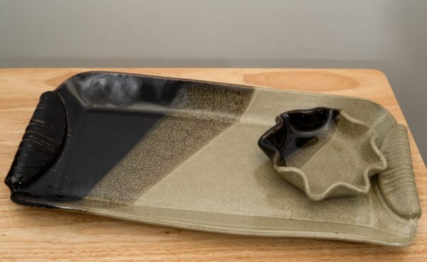 the photograph shows a clay coyote large tray and little dipper combination resting on a light colored wooden table. the large tray and little dipper are both glazed in mint chip. the little dipper is the circular or star style. both the large tray and the little dipper have the midnight black part of the glaze positioned to be pointing left. the little dipper is resting on the very right side of the large tray, with one of the points of the little dipper overlapping with the large trays right side handle. the large tray is at a slight angle relative to the tables positioning, with the left side pointed slightly higher, and the right side slightly lower.