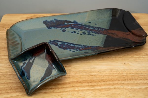 the photograph shows a clay coyote large tray and little dipper combo resting on a light colored wooden table. the large tray and little dipper are both glazed in zappa. the little dipper is the square style. it is resting on the lower left corner of the large tray, slightly lifted by the large trays lip, giving a good view of the glaze pattern inside the little dipper. the large tray is at a slight angle, with the left side slighting pointing down towards the lower left corner of the photo, and the right side pointed towards the upper right. the upper right corner of the large tray is just out of frame.