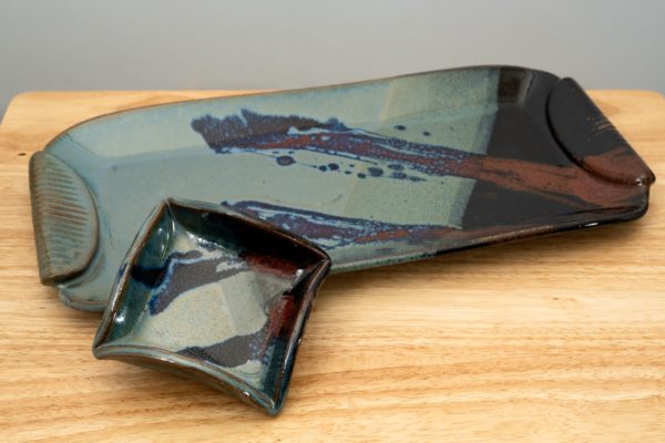 the photograph shows a clay coyote large tray and little dipper combination resting on a light colored wooden table. the little dipper and large tray are both glazed in zappa. the little dipper is the square style. it is resting on the lip of the large tray. it is resting near the lower left corner of the large tray. the little dipper is resting in a way to be slightly elevated to show the glaze patter on the bottom. the tray is resting at an angle. the trays left side is pointed slightly downwards and the right is pointed slightly upwards. the background is a white painted wall. the photograph is well lit with white light.