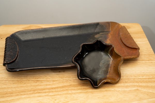 the photograph shows a clay coyote large tray and little dipper both resting on a light colored wooden table. the large tray and little dipper are both glazed in mocha swirl. the small brown section of the mocha swirl pattern is facing to the right on both pieces. the little dipper is the circular or star style of dipper. the little dipper is resting on the outside right lip of the tray, propping the little dipper up slightly to show off the glaze pattern within. the background is a white wall.