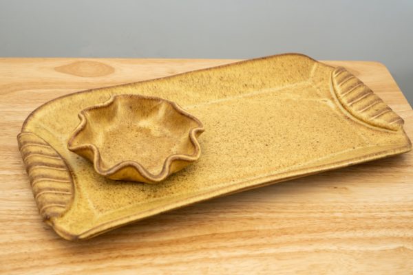 the photograph shows a clay coyote large tray and little dipper resting on a light colored wooden table. both clay coyote pieces are glazed in yellow salt. the little dipper is the circular or star style of dipper. the little dipper is resting on the tray on the left side of it. the tray is slightly angled compared to the table. the right side of the tray is pointing to the upper right corner, with the left side pointed towards the lower left corner. the background is a plain white wall.