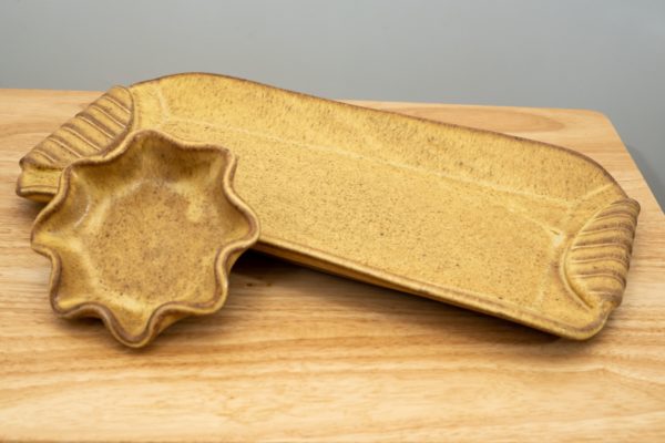 the photograph shows a clay coyote large tray and little dipper resting on a light colored wooden table. the large tray and the little dipper are both glazed in yellow salt. the little dipper is the circular or star pattern dipper. the little dipper is resting outside of the tray on the left side of the tray. the large tray is angled slightly in comparison to the table. the tray is pointed towards the upper left corner and lower right corner of the photograph. the background is a white wall.