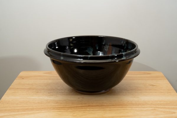 the photograph shows a clay coyote deep salad bowl glazed in midnight garden resting on a small light colored wooden table. the background is a white wall. the photograph is well lit with white light.