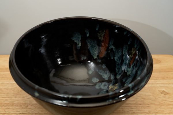 the photograph shows a high angle view of a clay coyote deep salad bowl glazed in midnight garden. the angle is high enough to allow the viewer to see the glaze details in the bowl. the deep salad bowl is resting on a small light colored wooden table. the background is a white wall.