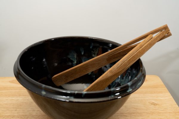 the photograph shows a clay coyote deep salad bowl glazed in midnight garden. It is resting on a small light colored wooden table. the deep salad bowl has a pair of baer woodworks wooden tongs resting on the right side of the bowl. the background is a white wall. the photograph is well lit with white light.