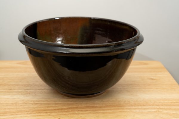 the photograph shows a clay coyote deep salad bowl glazed in merlot resting on a small light colored wooden table. the background is a white wall. the photograph is well lit with white light.