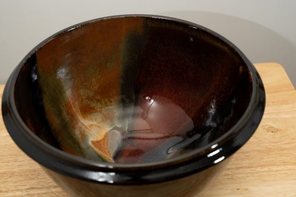 the photograph shows a high angle view of a clay coyote deep salad bowl glazed in merlot. the angle is high enough to allow the viewer to see the glaze details in the bowl. the deep salad bowl is resting on a small light colored wooden table. the background is a white wall.