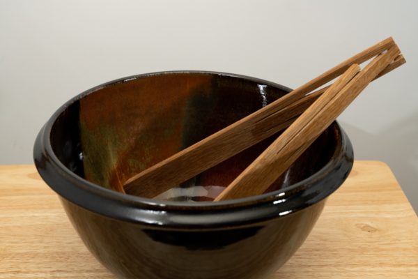 the photograph shows a clay coyote deep salad bowl glazed in merlot. It is resting on a small light colored wooden table. the deep salad bowl has a pair of baer woodworks wooden tongs resting on the right side of the bowl. the background is a white wall. the photograph is well lit with white light.