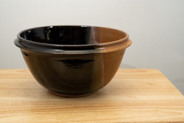 the photograph shows a clay coyote deep salad bowl glazed in mocha swirl resting on a small light colored wooden table. the background is a white wall. the photograph is well lit with white light.