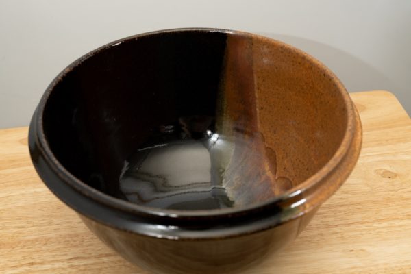 the photograph shows a high angle view of a clay coyote deep salad bowl glazed in mocha swirl. the angle is high enough to allow the viewer to see the glaze details in the bowl. the deep salad bowl is resting on a small light colored wooden table. the background is a white wall.