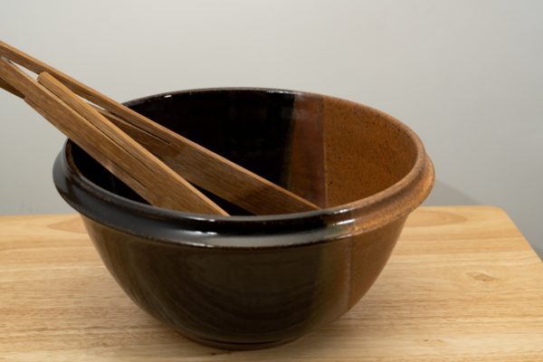 the photograph shows a clay coyote deep salad bowl glazed in mocha swirl. It is resting on a small light colored wooden table. the deep salad bowl has a pair of baer woodworks wooden tongs resting on the left side of the bowl. the background is a white wall. the photograph is well lit with white light.