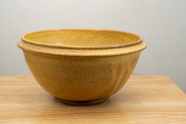 the photograph shows a clay coyote deep salad bowl glazed in yellow salt resting on a small light colored wooden table. the background is a white wall. the photograph is well lit with white light.