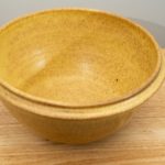 the photograph shows a high angle view of a clay coyote deep salad bowl glazed in yellow salt. the angle is high enough to allow the viewer to see the glaze details in the bowl. the deep salad bowl is resting on a small light colored wooden table. the background is a white wall.
