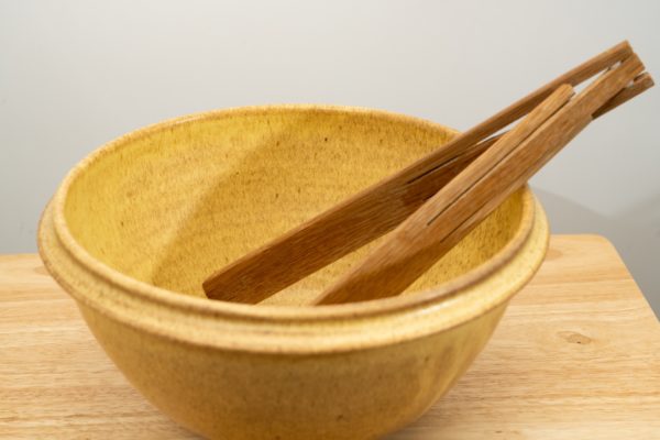 the photograph shows a clay coyote deep salad bowl glazed in yellow salt. It is resting on a small light colored wooden table. the deep salad bowl has a pair of baer woodworks wooden tongs resting on the right side of the bowl. the background is a white wall. the photograph is well lit with white light.