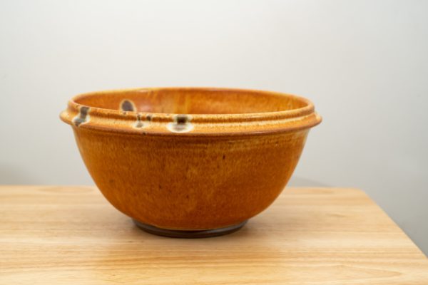 the photograph shows a clay coyote deep salad bowl glazed in feather resting on a small light colored wooden table. the background is a white wall. the photograph is well lit with white light.