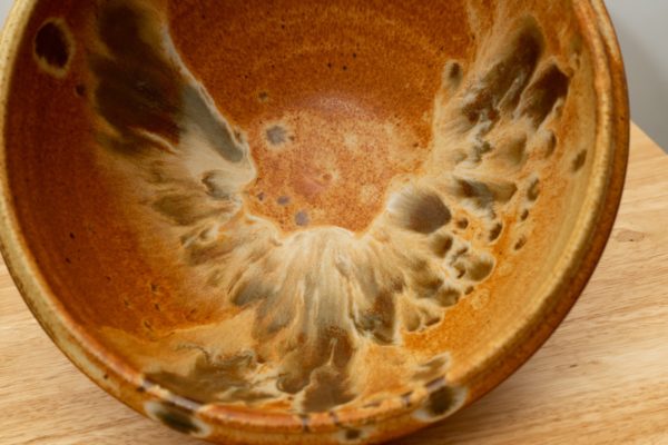 the photograph shows a clay coyote deep salad bowl that is glazed in feather. the bowl is on its side to show off the feather design on the inside bottom and walls of the bowl. the glaze looks like a bird in flight. the table it is resting on is a light wooden color, and the background is a white wall. the photograph is well lit with white light.