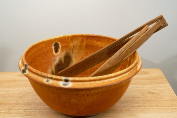 the photograph shows a clay coyote deep salad bowl glazed in feather. It is resting on a small light colored wooden table. the deep salad bowl has a pair of baer woodworks wooden tongs resting on the right side of the bowl. the background is a white wall. the photograph is well lit with white light.