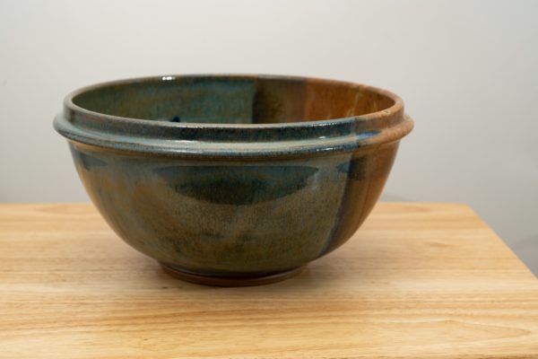 the photograph shows a clay coyote deep salad bowl glazed in joes blue resting on a small light colored wooden table. the background is a white wall. the photograph is lit with white light.