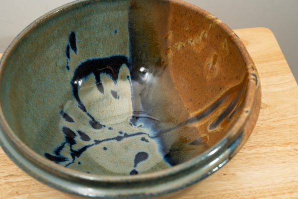 the photograph shows a high angle view of a clay coyote deep salad bowl glazed in joes blue. the angle is high enough to allow the viewer to see the glaze details in the bowl. the deep salad bowl is resting on a small light colored wooden table. the background is a white wall.