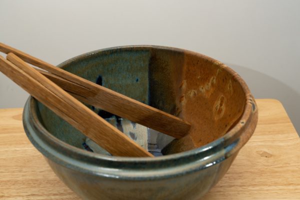 the photograph shows a clay coyote deep salad bowl glazed in joes blue. It is resting on a small light colored wooden table. the deep salad bowl has a pair of baer woodworks wooden tongs resting on the left side of the bowl. the background is a white wall. the photograph is lit with white light.