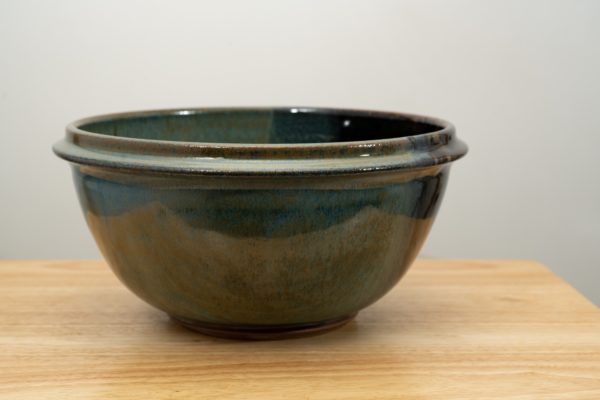 the photograph shows a clay coyote deep salad bowl glazed in zappa resting on a small light colored wooden table. the background is a white wall. the photograph is lit with white light.