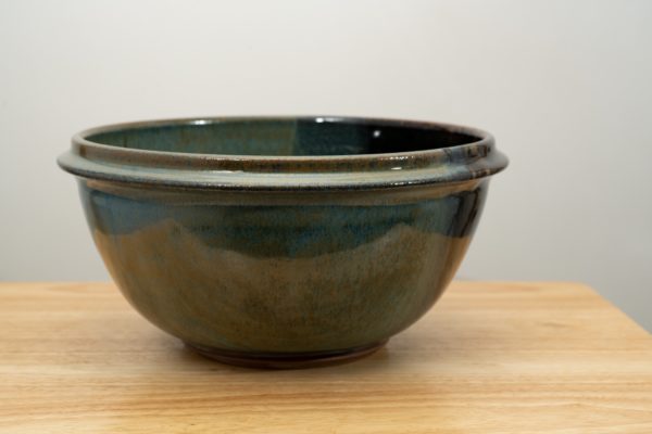 the photograph shows a clay coyote deep salad bowl glazed in zappa resting on a small light colored wooden table. the background is a white wall. the photograph is lit with white light.