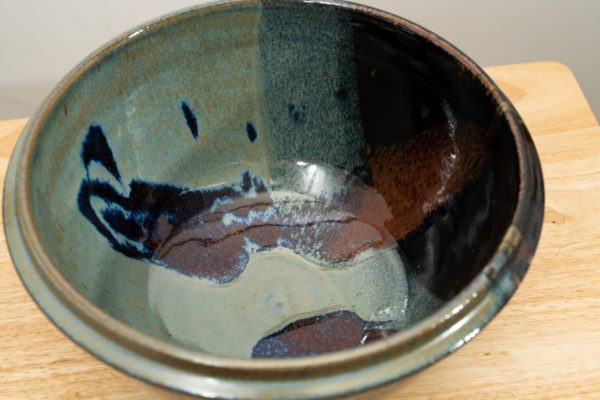 the photograph shows a high angle view of a clay coyote deep salad bowl glazed in zappa. the angle is high enough to allow the viewer to see the glaze details in the bowl. the deep salad bowl is resting on a small light colored wooden table. the background is a white wall.