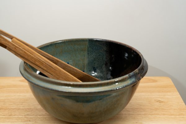 the photograph shows a clay coyote deep salad bowl glazed in zappa. It is resting on a small light colored wooden table. the deep salad bowl has a pair of baer woodworks wooden tongs resting on the left side of the bowl. the background is a white wall. the photograph is lit with white light.