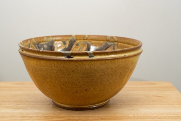 the photograph shows a clay coyote deep salad bowl glazed in tequila sunrise resting on a small light colored wooden table. the background is a white wall. the photograph is well lit with white light.