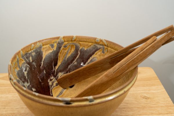 the photograph shows a clay coyote deep salad bowl glazed in tequila sunrise. It is resting on a small light colored wooden table. the deep salad bowl has a pair of baer woodworks wooden tongs resting on the right side of the bowl. the background is a white wall. the photograph is lit with white light.