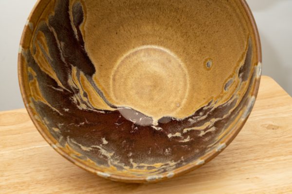 the photograph shows a clay coyote deep salad bowl glazed in tequila sunrise resting on a small light colored wooden table. the bowl is resting on its side to provide a clear view of the glaze pattern on the inside of the bowl. the background is a white wall. the photograph is lit with white light.