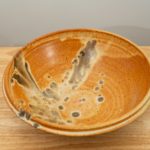 the photograph shows a high angle view of a clay coyote shallow salad bowl resting on a light colored small wooden table. the angle allows the view to see the feather pattern on the inside of the bowl. the background is a white wall. the photograph is well lit with white light.