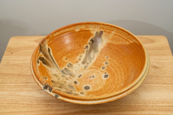 the photograph shows a high angle view of a clay coyote shallow salad bowl resting on a light colored small wooden table. the angle allows the view to see the feather pattern on the inside of the bowl. the background is a white wall. the photograph is well lit with white light.