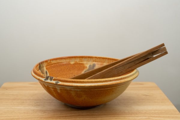 the photograph shows a clay coyote shallow salad bowl with a pair of baer woodworks foldable salad tongs resting in them. the shallow salad bowl is glazed in feather. the shallow salad bowl is sitting on a small light colored wooden table. the baer woodworks tongs are resting on the right side rim of the shallow salad bowl. the tongs are made of dark wood. the background is a white wall.